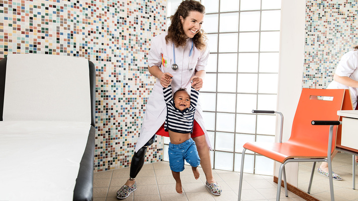 Standing with her legs wide apart, Marije playfully swings a young patient around by her hands in a treatment room.