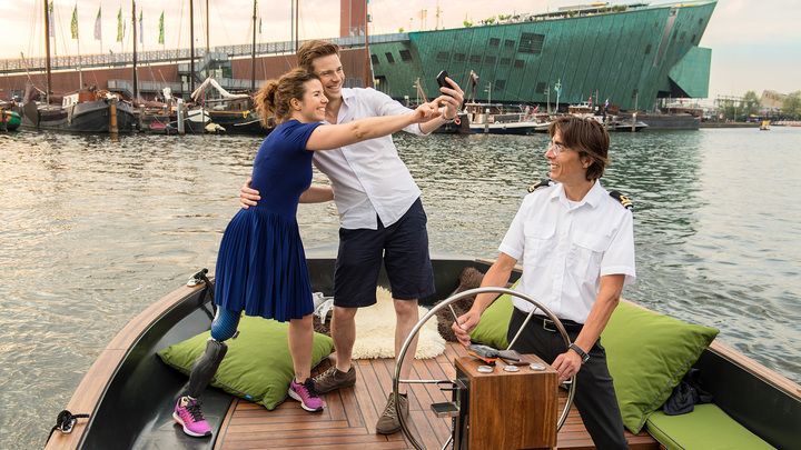 Marije und ihr Freund stehen am Bug eines Touristenbootes und machen Selfies mit dem Smartphone.