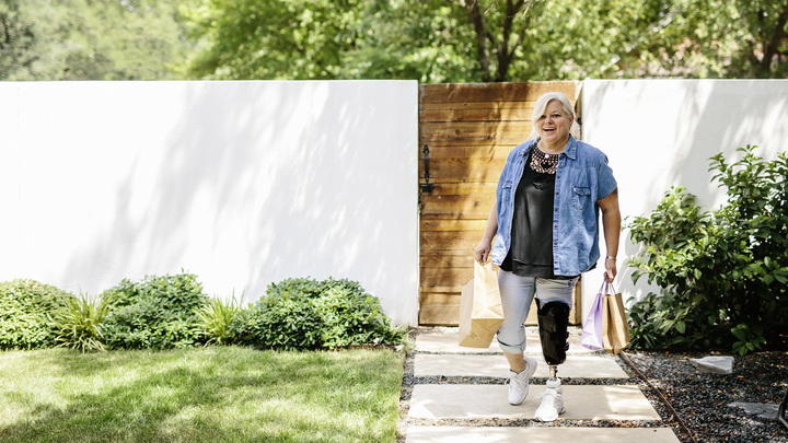 Diana wears her Meridium prosthetic foot as she walks up the uneven path to her house with shopping bags.
