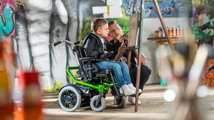 Charly, Berlin yeşil Ottobock elektrikli tekerlekli sandalyesi Skippi üzerinde resim sehpasında çizim yaparken.